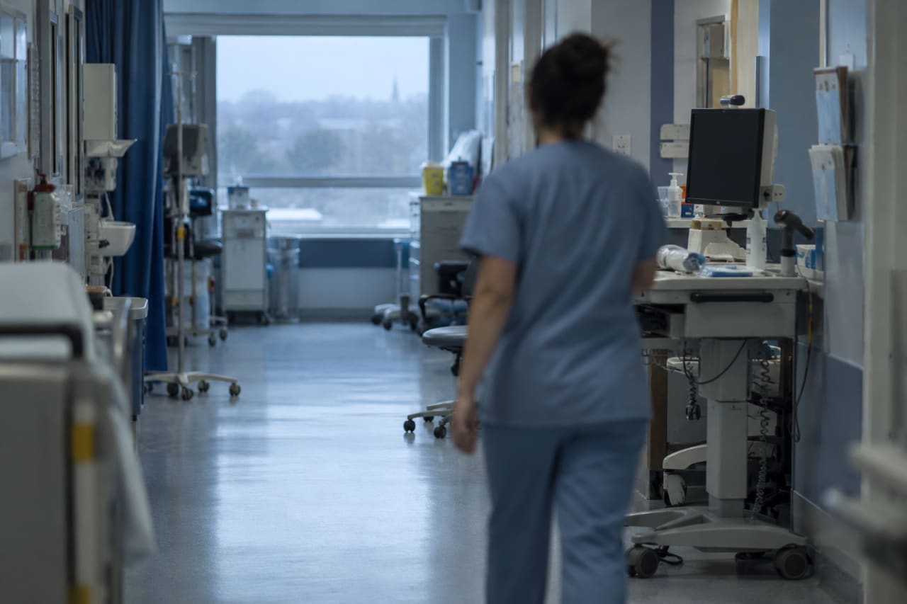 A clinician in blue scrubs, seen from behind, walking down a hospital ward corridor past a computer workstation cart
